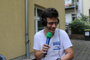 Im Bild ist Redaktionsmitglied Mirko. Er trägt ein weißes T-Shirt mit dem blauen Aufdruck Radio Inklusive. In der Hand hält er ein blau-grünes Mikrofon. Auf den Ohren hat Mirko große Kopfhörer. Er lacht und spricht in das Mikrofon. Im Hintergrund befindet sich ein Hinterhof mit Mülltonne und Büschen.