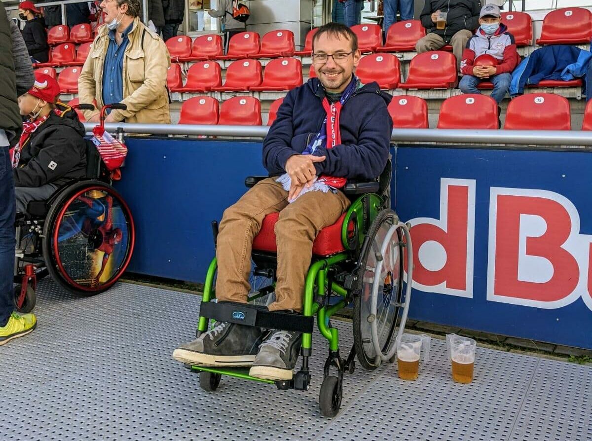 David sitz auf der Rollstuhltribüne im Stadion von RB Leipzig und grinst frontal in die Kamera. Er sitzt in einem grünen Rollstuhl neben dem zwei Becher mit Bier stehen. Davids trägt eine hellbraune Hose und eine dunkle jacke. Die Sonne scheint.