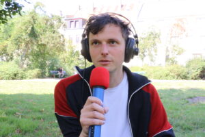 Erik Tenzler sitzt auf einer Wiese in einem Park. Er trägt eine rot-schwarze Trainingsjacke und ein weißes T-Shirt. Er hat große Kopfhörer auf. In der rechten Hand hält er ein blau-rotes Mikrofon. Er schaut nicht in die Kamera und spricht in das Mikrofon. Im Hintergrund sind verschwommen Bänke, Bäume, Büsche und Häuser zu sehen.