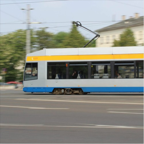 Zu sehen ist eine Straßenbahn in Leipzig. Sie ist grau und hat einen gelben und einen blauen Streifen. Im Vordergrund ist ein Stück Straße. Im Hintergrund ein Haus, grüne Baumkronen und ein grauer Strommast.