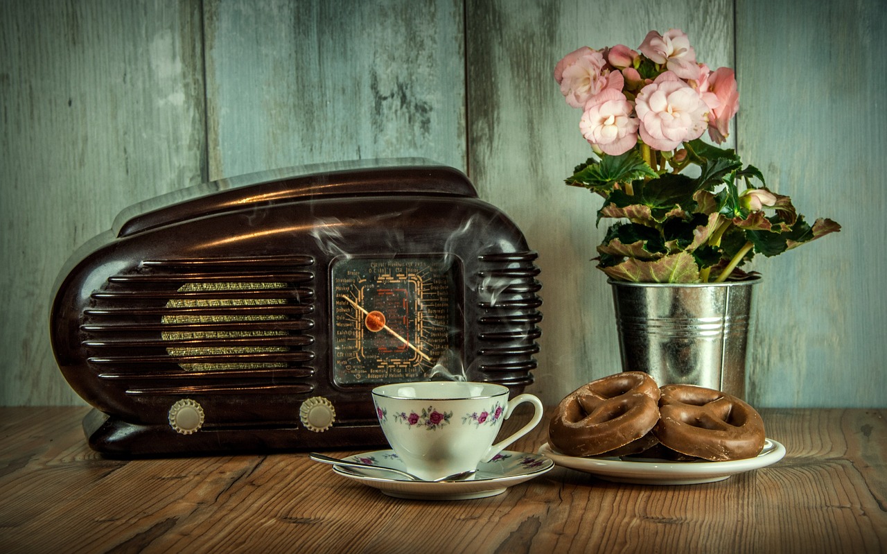 Ein altes Radio steht auf einem Tisch. Daneben eine Kaffeetasse in Retro-Optik, ein Teller mit Lebkuchen-Brezeln und eine Blume im Topf. Auf dem gesamten Bild liegt ein Foto-Filter, der das Bild alt wirken lässt.