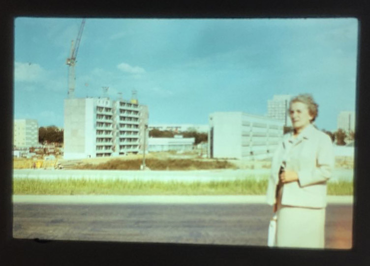 Eine alte grauhaarige Frau in weißen Kostüm. Der Wind zerzaust ihr die Haare. Ihr Gesichtsausdruck ist ernst. Im Hintergrund werden die Plattenbauten im Leipziger Stadtteil Grünau errichtet.