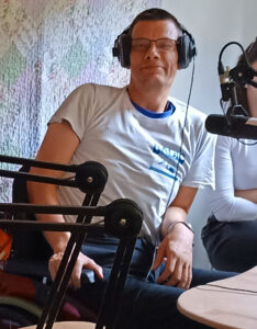 Redaktions-Mitglied Mirko. Er trägt ein weißes Shirt mit dem Radio-Inklusive-Logo. Das Logo ist blau und stellt ein Radio und ein Mirkofon dar - kombiniert mit dem Schriftzug "Radio Inklusive". Mirko hat Kopfhörer auf. Vor ihm ist ein Mirkofon aufgebaut. Mirko lächelt in die Kamera.