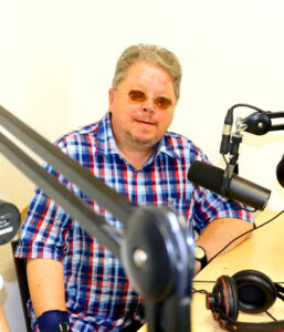 Redakteur Uwe. Er sitzt im Tonstudio am Mikrofon und lächelt in die Kamera. Er trägt ein kariertes Hemd und eine Sonnenbrille.