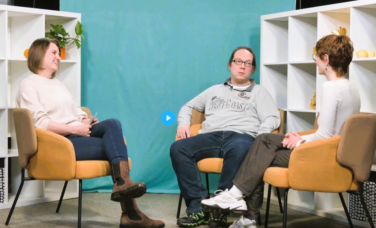 Die Redaktionsmitglieder Johannes und Adele im Interview mit einer Moderatorin. Johannes und Adele tragen jeweils eine Brille, ein helles Oberteil, dunkle Hosen und Turnschuhe. Ebenso die Moderatorin. Sie sitzen in gelben Stühlen und unterhalten sich angeregt. Im Hintergrund sind weiße Regale mit Pflanzen zu sehen. Dahinter ist eine türkisfarbene Wand.