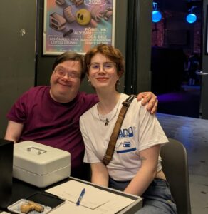 Redakteurin Adele. Sie trägt ein weißes T-Shirt mit der Aufschrift Radio Inklusive. Sie lächelt und trägt eine Brille. Sie ist jung. Neben ihr sitzt Redakteur Steffen. Auch er lächelt und hat seinen Arm um Adele gelegt.