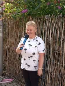 Redakteurin Antje im sommerlichen Outfit, mit weißem T-Shirt mit Muster. Sie hat blonde kurze Haare und lächelt in die Kamera. Sie spricht in ein Mikrofon, das sie in der Hand hält. Im Hintergrund ein Zaun und Blumen.