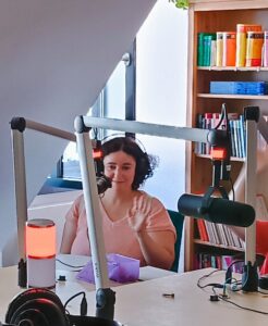 Redakteurin Elvira in einem Radiostudio. Sie hat langes, lockiges, dunkles Haar und ein lachsfarbenes Oberteil an. Sie lächelt und winkt in die Kamera.