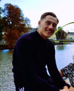 Redakteur und Bundesfreiwilligendienstler Oskar. Er sitzt auf einem Geländer an einem Fluss. Er trägt einen dunkelblauen Pullover, ist jung, trägt einen Schnauzbart und hat einen Seitenscheitel. Er lächelt in die Kamera.