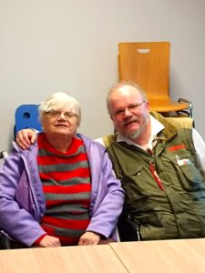 Redakteurin Edith und Redakteuer Alexander sitzen an einem Tisch. Edith ist 72 Jahre alt, hat weißes, kurzes Haar und eine Brille. Sie trägt winterliche Kleidung. Alexander hat eine Halbglatze und einen Bart sowie eine Brille. Auch er ist warm angezogen. Beide lächeln und Alexander hat seinen Arm um Edith gelegt.