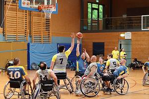 Etwa 7 Personen spielen in einer Turnhalle Rollstuhlbasket. Ein Spieler wirft gerade den Ball Richtung Korb. Ein anderer versucht, den Wurf zu blocken.