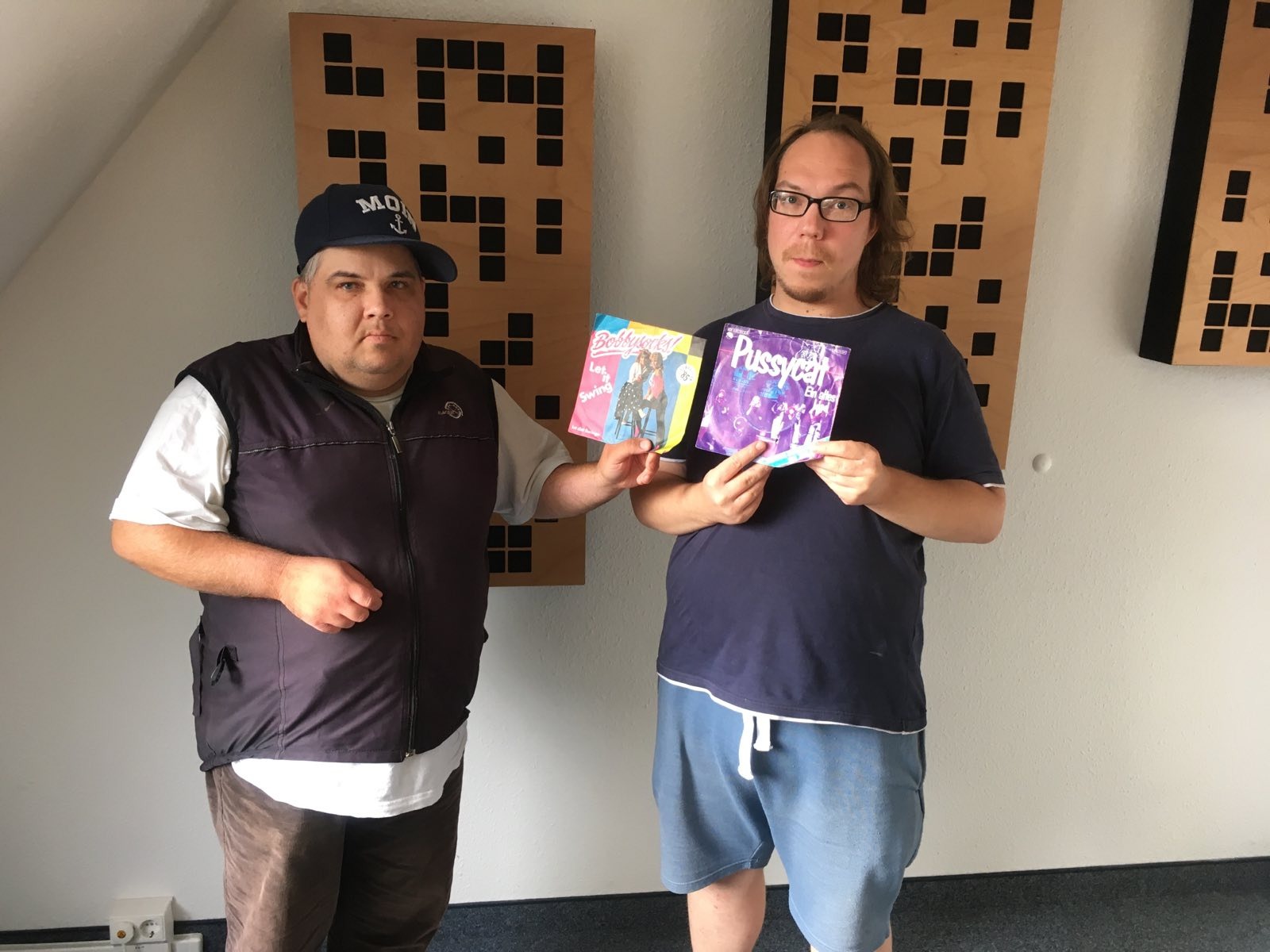 Die Radio-DJs Mütze und Johannes. Sie tragen sommerliche Kleidung und halten ihre Lieblingsschallplatten hoch. Im Hintergrund hängen Akustikelemente eines Tonstudios an einer weißen Wand.