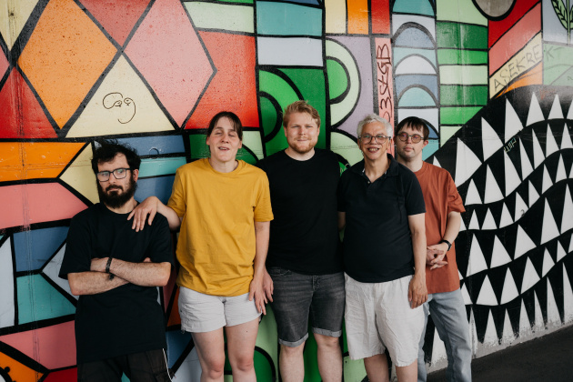 5 Personen der Gruppe Blumenstrauss. 3 Männer, zwei Frauen. Sie tragen dunkle, sommerliche Kleidung und schauen ernst in die Kamera. Im Hintergrund eine bunte Wand.