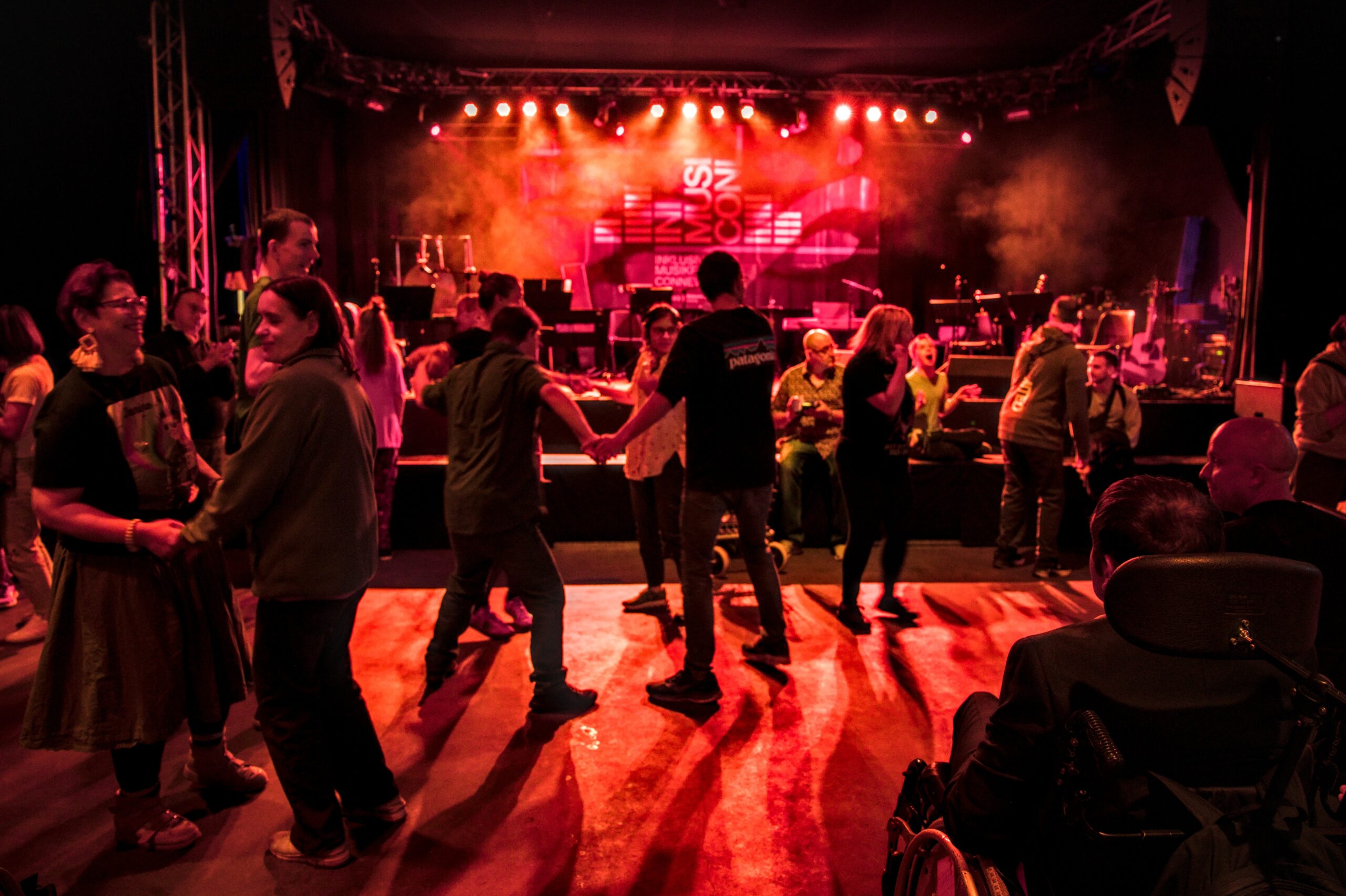 Ein Disko-Raum in rotem Licht. Viele Leute tanzen. Im Hintergrund ist eine Bühne ebenfalls in rotes Licht getaucht. An der Rückwand ein Plakat: INMUSICON.