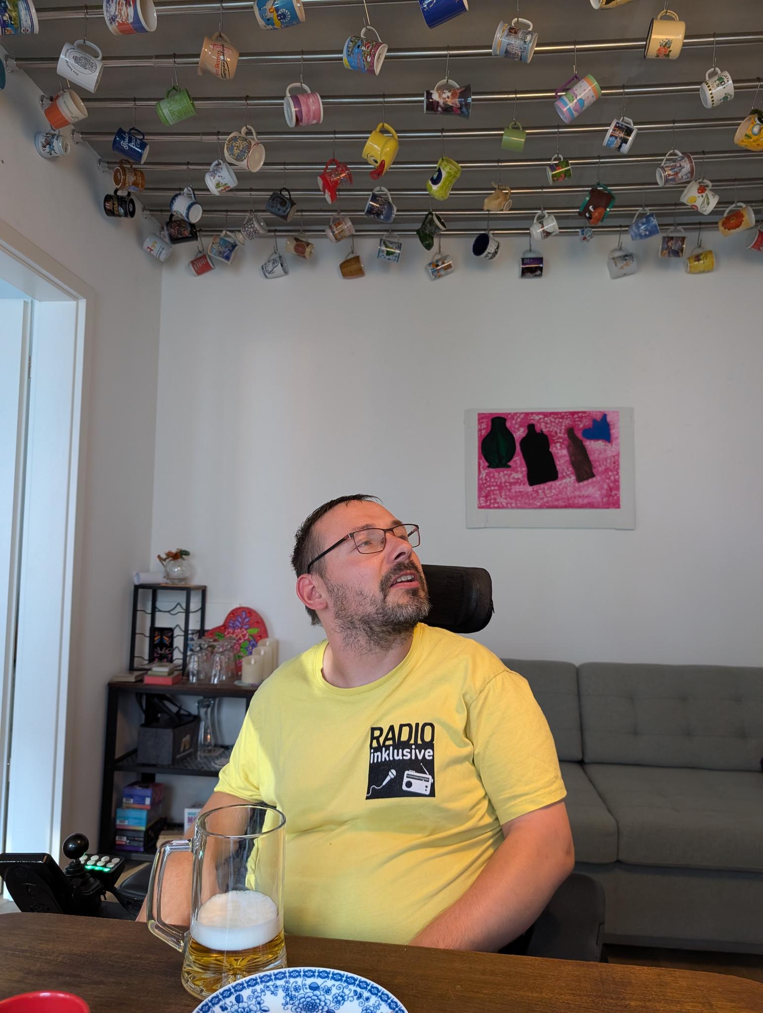 Radio Inklusive Redakteur Davind in seinem Wohnzimmer. Er sitzt in seinem Rollstuhl am Wohnzimmertisch. Er spricht angeregt und trägt ein gelbes T-Shirt. An der Decke hängt seine Tassensammlung. Im Hintergrund steht ein graues Sofa.